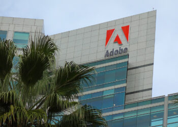 The image shows a modern, multi-story office building with the Adobe Stock logo and name prominently displayed near the top. The building is surrounded by palm trees, and the sky is cloudy. | FintechZoom