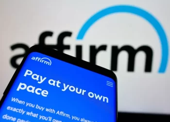 A smartphone screen displaying the Affirm app with the text "Pay at your own pace" is in focus, while the blurred Affirm logo and Affirm Stock chart appear in the background on a larger screen. | FintechZoom