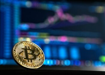 A close-up of a gold Bitcoin coin in sharp focus, positioned on a dark surface. The background is blurred and features a computer screen displaying financial charts and data with various colors and lines indicating market trends, reminiscent of KuCoin's detailed analytics. | FintechZoom