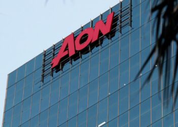 Close-up of a modern glass high-rise building with the red Aon logo prominently displayed at the top. The blue-tinted windows reflect the sky, while some greenery is partially visible in the foreground—a striking image for anyone tracking Aon stock. | FintechZoom