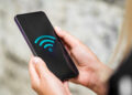 Close-up of a person holding a smartphone with an LPB Piso Wifi symbol displayed on the screen. The background is blurred, and the person’s hands are visible, holding the phone gently. | FintechZoom
