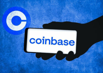 A silhouette of a hand holds a smartphone displaying the word "coinbase" on the screen against a blue textured background. To the left, the Coinbase logo, a white "C" inside a blue circle, is prominently visible, symbolizing Coinbase's presence in the stock market. | FintechZoom