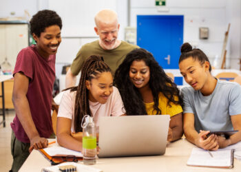 A diverse group of five people, consisting of four young adults and one older adult, gather around a laptop in a bright room. They are smiling and focused, suggesting collaborative work or learning. Notebooks, a tablet, and a water bottle are on the table as they discuss ways to gamify your classroom. | FintechZoom