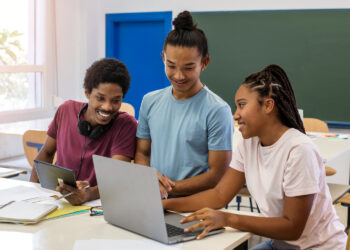 Three students are working together at a table in a classroom. One person on the left is holding a tablet, the person in the middle is pointing at the laptop held by the person on the right. They appear engaged and friendly, possibly exploring game-based learning platforms, with notebooks and papers scattered on the table. | FintechZoom