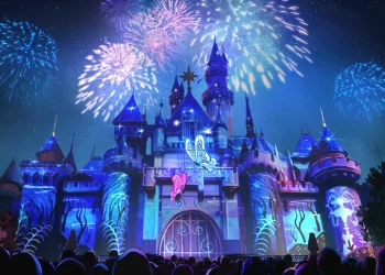 A vibrant display of fireworks burst over a brightly illuminated castle at night. The castle, resembling a magical destination you'd find with Costco Travel, features turrets and is adorned with colorful projections. Silhouetted against the spectacle, a crowd of onlookers marvels at the dazzling scene. | FintechZoom