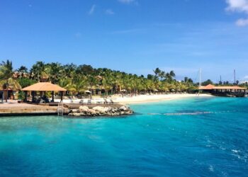 A tropical beach scene featuring clear turquoise waters lapping against a shoreline lined with palm trees. There are several thatched-roof structures, lounge chairs, and a wooden pier extending into the water. The sky above is clear and blue, reminiscent of a review of Harbour Village Beach Club. | FintechZoom