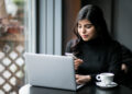 A woman with long dark hair sits at a table in a cafe, using her phone while working on a laptop, perhaps browsing VNSGU Net for her research. She is wearing a black top and is focused on her devices. A cup of coffee is placed on the table beside her. The background shows a window with blurred outside view. | FintechZoom