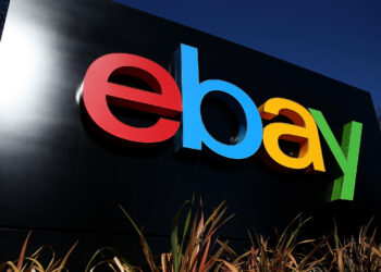 A large outdoor sign proudly displays the eBay logo, with "e" in red, "b" in blue, "a" in yellow, and "y" in green against a black background. The striking sign is surrounded by tall plants and set against a clear blue sky. | FintechZoom