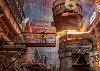 A worker in protective gear stands on a platform beside a large, industrial furnace pouring molten metal. Sparks fly as the bright, glowing liquid flows from the furnace. The background shows the interior of a spacious factory with US Steel Stock steel beams and machinery. | FintechZoom