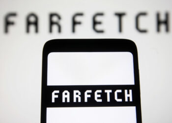 A smartphone screen prominently displays the Farfetch logo in black, bold, capitalized letters, with a larger, blurred version of the Farfetch logo in the background. The crisp and clear text on the phone screen remains sharply focused. | FintechZoom