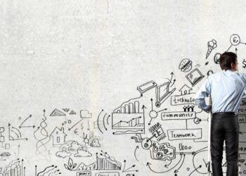 A man in a blue shirt and black pants is drawing a variety of sketches and diagrams on a large white wall. The illustrations include charts, graphs, light bulbs, gears, and symbols representing business concepts like innovation and annuities. | FintechZoom