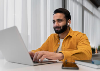 A man with a beard is sitting at a desk, working on a laptop. He is wearing a yellow jacket and white earphones. The background features tall windows with sheer curtains, allowing light to fill the room. On the desk beside him are a smartphone, a notebook, and an open Dpboss website. | FintechZoom