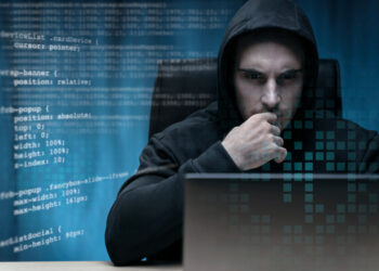 A person in a hooded sweatshirt sits at a desk, focused intently on a laptop. The background features various lines of code and a digital interface overlay, emphasizing the theme of cybersecurity or hacking, particularly Denial-of-Service Attacks. The environment is dimly lit, creating a serious atmosphere. | FintechZoom