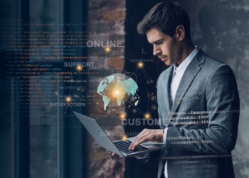 A man in a gray suit uses a laptop, standing against a background of digital graphics featuring code, a floating globe, and words like "ONLINE," "SUPPORT," "CUSTOMER," and "COMMUNICATION." The scene highlights the fusion of technology, business, and essential cybersecurity tips. | FintechZoom