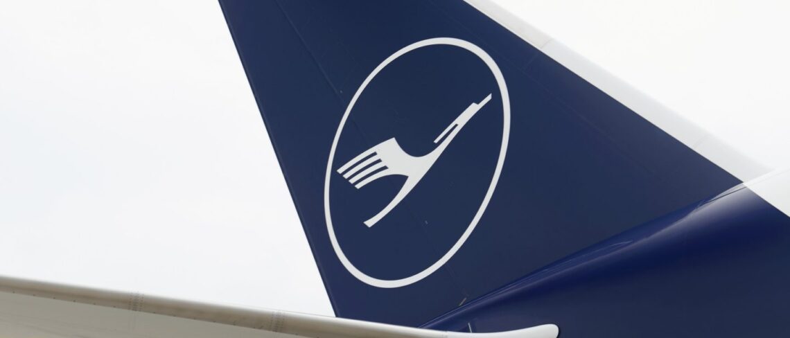 A close-up of the tail of an aircraft featuring the Lufthansa logo, with a white silhouette of a crane in flight inside a white circle, set against a dark blue background. The sleek design is evident whether it's on Airbus stock or Boeing stock. | FintechZoom