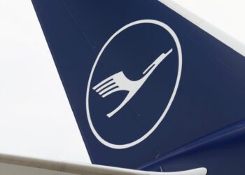 A close-up of the tail of an aircraft featuring the Lufthansa logo, with a white silhouette of a crane in flight inside a white circle, set against a dark blue background. The sleek design is evident whether it's on Airbus stock or Boeing stock. | FintechZoom