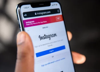 A person holds a smartphone displaying the Instagram log-in screen. The phone screen shows the URL instagram.com at the top, with a banner suggesting to download the app for features like Story Saver. The device is held in the person's right hand with the thumb visible on the left side. | FintechZoom
