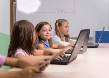 A group of young children is seated at a desk in a classroom, each using a laptop. The focus is on a smiling girl in the middle, with two other girls working on laptops on either side, likely interacting with Blooket. A whiteboard is visible in the background. | FintechZoom