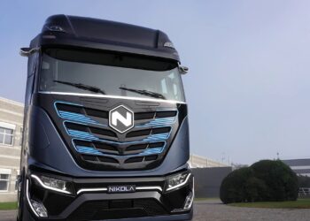 A black Nikola electric semi-truck is parked outdoors in a modern industrial setting, highlighting blue accents and the Nikola logo at the front. Buildings and greenery set a contrasting backdrop, reflecting the innovative spirit that drives interest in Nikola stock. | FintechZoom