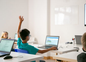 A group of students sitting at desks in a bright, modern classroom with laptops open. One student, wearing glasses and a blue shirt, is raising his hand. The whiteboard stands ready for new ideas, and in the background, another student enjoys their gamified learning experience. | FintechZoom