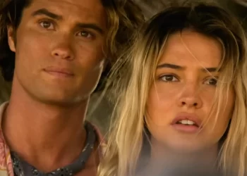 Close-up of a young man and woman, reminiscent of the Outer Banks cast, standing next to each other, looking slightly off to the side with serious expressions. The man has wavy hair, wears a shirt, and has a cloth around his neck. The woman has long, tousled hair and an open-mouthed expression. | FintechZoom
