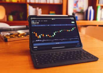 A tablet with a detachable keyboard displaying a financial chart with candlestick patterns sits on a wooden table. In the background, there are bookshelves, a plant, and a tray with coins. As the cornerstone of this casual home workspace, it seamlessly blends function and style. | FintechZoom
