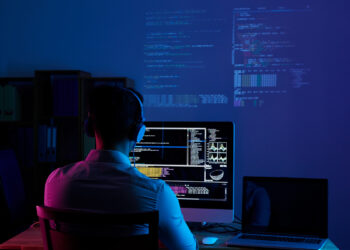 A person wearing headphones is sitting at a desk, facing a computer monitor displaying multiple lines of code and various data visualizations. There is a laptop beside them showing details on malware attacks, with more code projected on the wall in the dimly lit room. | FintechZoom
