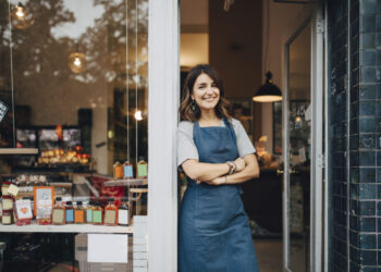 A smiling person stands at the entrance of a store, wearing a blue apron over a light gray shirt. Their arms are crossed, and the store's shelves, filled with various jars and products, are visible in the background. The atmosphere is warm and welcoming—an ideal example of how to write a business plan for a small business. | FintechZoom