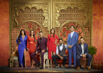 A group of elegantly dressed individuals pose in front of an ornate golden backdrop with intricate designs. Four women and three men stand and sit in various stylish outfits, exuding confidence and unity as though attending a grand book show in a richly decorated room. | FintechZoom