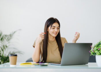 A woman with long brown hair, wearing a beige shirt, is sitting at a desk and looking at a gray laptop. A coffee cup, a phone, and a notebook are on the desk, and a potted plant is visible in the background. She appears to be engaging in a video call while browsing Movierulz. | FintechZoom