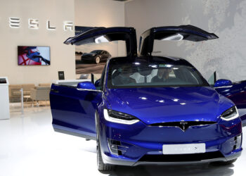 A blue Tesla Model X is displayed in a showroom with its signature falcon-wing doors open. In the background, a Tesla logo on the wall, a seating area, and a map create an inviting atmosphere. The sleek, modern design of the showroom mirrors the performance that has driven Tesla stock to new heights. | FintechZoom
