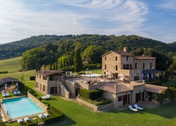 A picturesque villa with stone architecture situated amidst lush greenery, this luxury property in Italy features multiple levels with spacious terraces, an outdoor dining area, and a large pool surrounded by lounge chairs. Rolling hills and dense trees create a serene backdrop. | FintechZoom
