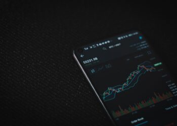 A smartphone screen displays a commission-free financial trading app with a candlestick chart, various data points, and indicators against a dark background. The chart shows upward and downward trends, with the price at 55231.98. | FintechZoom