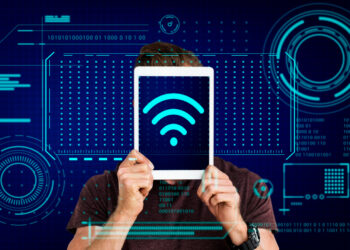 A person holds up a tablet with a blue Wi-Fi signal icon on the screen, covering their face. The background features a futuristic digital interface with holographic elements and binary code, reminiscent of a Pisonet Wifi Machine, creating a tech-themed visual. | FintechZoom
