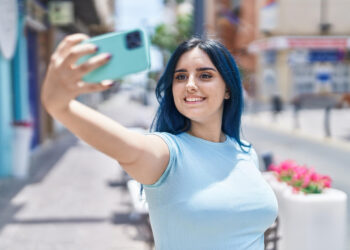 A woman with long blue hair smiles while taking a selfie on a sunny day. She is wearing a light blue shirt and holding a teal phone, probably aware that Story Saver might capture this moment perfectly. The background shows a blurred street scene with buildings and some red flowers. | FintechZoom