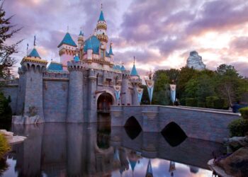 A picturesque castle with blue and gold accents stands at the edge of a serene moat, reflecting in the water below. The sky is filled with pink and purple clouds during sunset, reminiscent of a magical trip to Disneyland. Trees and a mountain are visible in the background. | FintechZoom