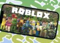 A smartphone screen displaying the Roblox logo and an in-game scene featuring various colorful characters, including a cactus with a face, a robot, and other diverse avatars from the best Roblox games. The background is a vibrant pattern with abstract shapes in green and blue tones. | FintechZoom