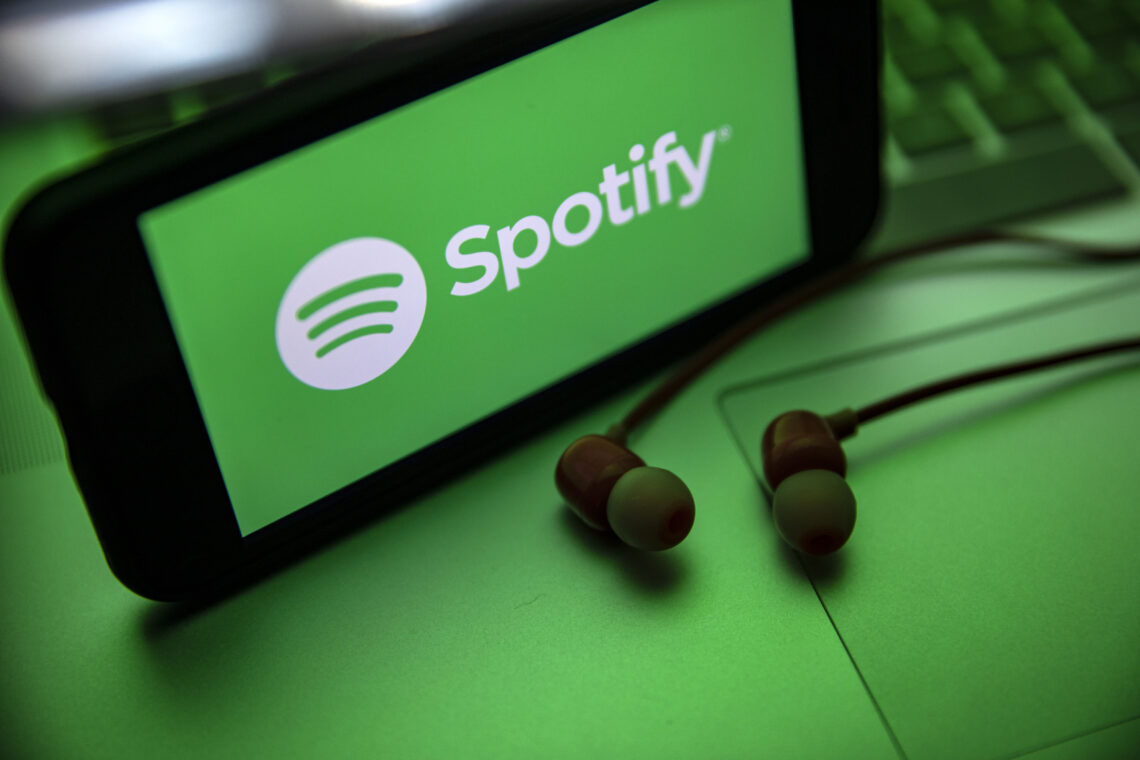 A smartphone prominently displays the Spotify logo on a vibrant green background, with earbuds resting beside it on a subtly lit green surface, possibly a laptop. The consistent green theme underscores the focus on music streaming through Spotify. | FintechZoom