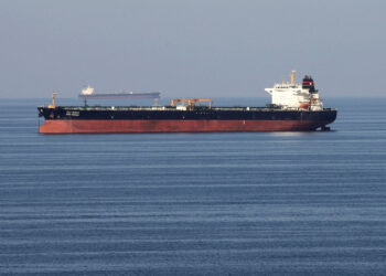 A large cargo ship with a red and black hull is sailing on calm blue waters under a clear sky, its journey reflecting the ebb and flow of oil prices. Another ship is visible in the distance on the horizon. | FintechZoom