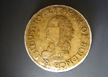 A brass-colored medallion with a profile of a person's head facing left in the center. Around the edge are the words "LIB. PAR. OF SCIENCE & INDUSTRY" and the year "1792" is inscribed below the profile image. The background is dark grey, resembling high Coin Grades in its meticulous detailing. | FintechZoom