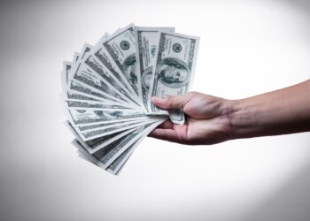 A hand is holding a fanned-out stack of one hundred dollar bills against a plain, softly lit background. The money is spread out, clearly showing Benjamin Franklin's portrait on each note, indicating a significant amount of money. The display of money exudes wealth and financial success. | FintechZoom