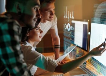 Three people focus on a computer screen displaying data visualizations. One person gestures towards the screen with a pen, explaining something, while the other two observe intently. The scene is well-lit, highlighting their engaged expressions as if they are analyzing trends for an online casino. | FintechZoom