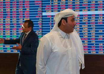 Two men in business attire stand in front of a large electronic display board showing stock market data with various numbers in red and blue. The man in the foreground, wearing traditional Middle Eastern clothing, discusses Sharia-compliant cryptocurrencies while the man in the background gestures animatedly. | FintechZoom