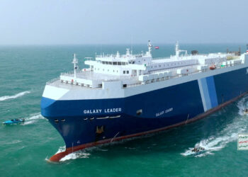 A large blue and white cargo ship named "Galaxy Leader" sails on the open sea. The ship features multiple decks and a flat top. Several smaller boats can be seen nearby, and the ocean extends to the horizon under a clear sky in the Red Sea. There is a sign in Arabic to the right. | FintechZoom