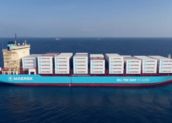 A large Maersk cargo ship painted in blue sails on a calm sea, carrying numerous white containers stacked neatly. The ship's side reads "MAERSK" and "ALL THE WAY TO ZERO." The clear sky above hints at the Maersk stock navigating towards a promising horizon. | FintechZoom