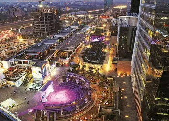 An evening aerial view of a modern cityscape featuring the illuminated commercial area of Cyber Hub Gurgaon. Buildings with glass facades and bustling streets are visible. A circular amphitheater-like structure with purple lights, ongoing construction, and busy roads are seen.
 | FintechZoom