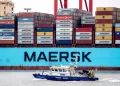 A large cargo ship with multiple Maersk shipping containers stacked on deck is docked at a port, notably as oil prices climb. In the foreground, a smaller coast guard boat is sailing by, flying a German flag. | FintechZoom