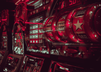 Close-up of brightly lit slot machines in a neon-lit casino. The spinning reels display "BAR" and star symbols, showcasing popular slot games. The surroundings are bathed in a red glow, emphasizing the captivating and energetic atmosphere of the gaming area. | FintechZoom