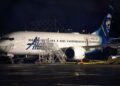 A commercial Boeing aircraft is parked on a wet tarmac at night. The plane, displaying the Alaska Airlines logo, is situated near portable stairs and ground support equipment. Dim lights can be seen in the background, along with a partial view of a vehicle. | FintechZoom