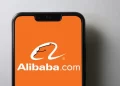 A smartphone with an orange screen displays the Alibaba.com logo, featuring a stylized 'A' with a smile and the text "Alibaba.com" in white. The phone is set against a plain light grey background, subtly hinting at the promising trends of Alibaba stock. | FintechZoom